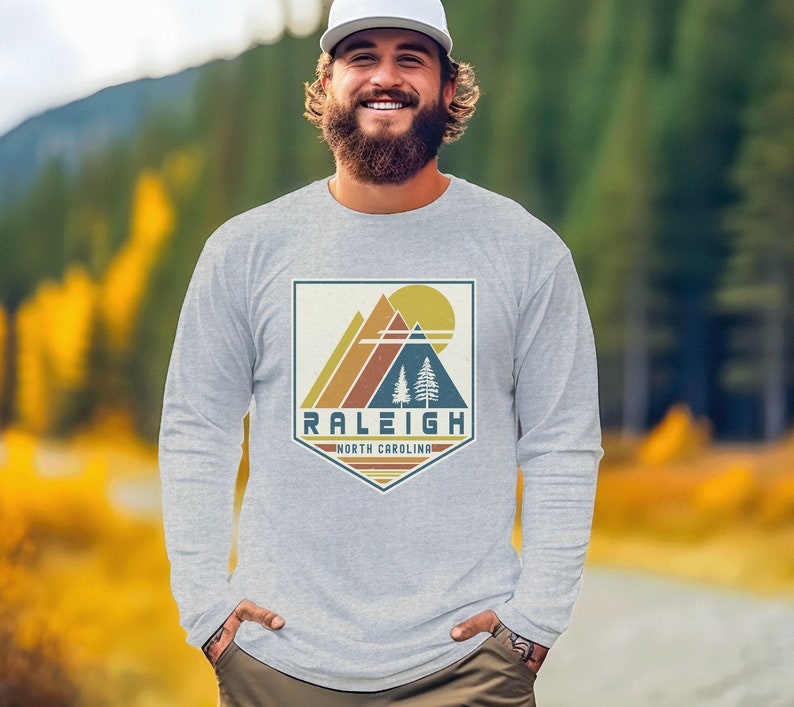 May include: A heather grey long-sleeve t-shirt with a vintage-style graphic of mountains, a sun, and trees. The graphic is in a shield shape with the text "Raleigh, North Carolina" below it.