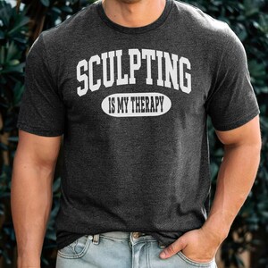 K&ouml;nnte beinhalten: Dunkelgraues T-Shirt mit dem Text "SCULPTING IS MY THERAPY" in wei&szlig;en Blockbuchstaben. Das Shirt hat einen Rundhalsausschnitt und kurze &Auml;rmel. Das Model tr&auml;gt eine blaue Jeans.