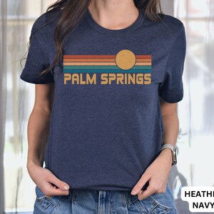 May include: A navy blue t-shirt with a retro sunset graphic and the text "PALM SPRINGS" in yellow. The graphic features a yellow sun with orange, red, teal, and brown stripes.