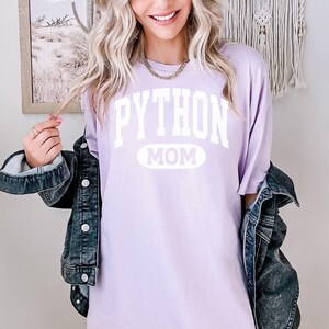 May include: A lilac t-shirt with the white text "PYTHON MOM". The shirt is paired with a denim jacket. The image suggests a relaxed, everyday style.