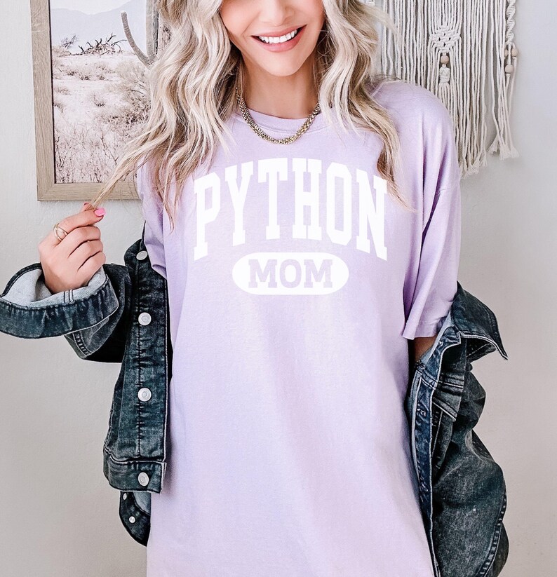 May include: A lilac t-shirt with the white text "PYTHON MOM". The shirt is paired with a denim jacket. The image suggests a relaxed, everyday style.