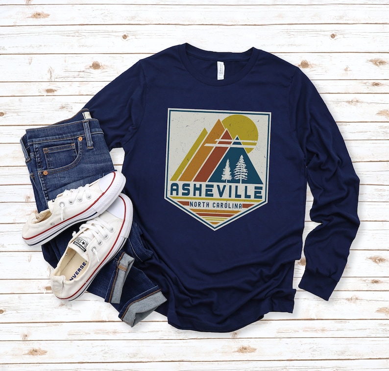 May include: Navy blue long-sleeve shirt with a vintage-style graphic of mountains, a sun, and trees. The graphic is in a faded, distressed style and includes the text "Asheville, North Carolina".