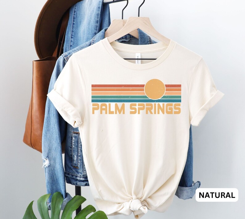 May include: A white t-shirt with a retro-style graphic design featuring a sunset with a sun and stripes in shades of orange, red, teal, and blue. The text "PALM SPRINGS" is printed in black below the graphic. The shirt is shown on a hanger with a denim jacket and a brown leather bag in the background.