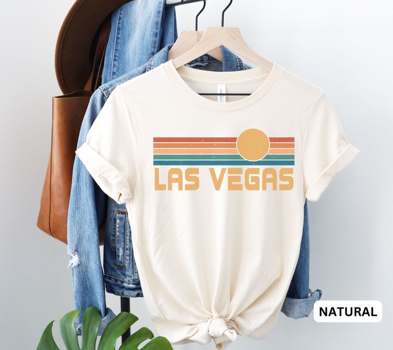 May include: A white t-shirt with a retro-style graphic of a sunset over a striped background with the text "LAS VEGAS". The shirt is displayed on a hanger with a denim jacket and a brown leather bag.