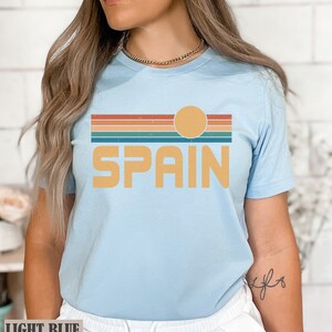 May include: Light blue t-shirt with a retro sunset graphic and the word "SPAIN" in yellow text.