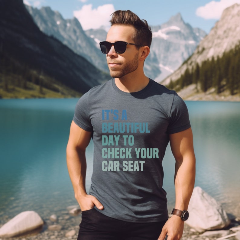 May include: A dark blue t-shirt with a white graphic that reads "It's a beautiful day to check your car seat". The shirt is being worn by a person standing in front of a lake and mountains.