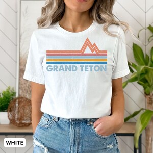 Puede incluir: Camiseta blanca con un gr&aacute;fico retro de monta&ntilde;as y el texto "GRAND TETON" en un dise&ntilde;o a rayas de colores.