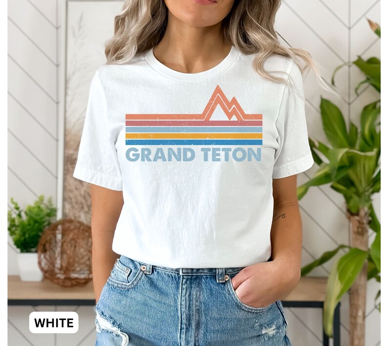 Puede incluir: Camiseta blanca con un gr&aacute;fico retro de monta&ntilde;as y el texto "GRAND TETON" en un dise&ntilde;o a rayas de colores.