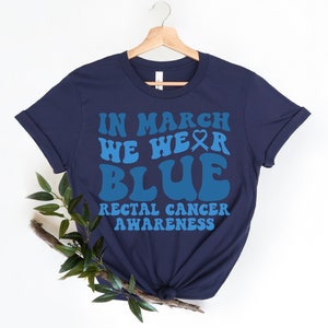 Puede incluir: Camiseta azul marino con el texto "In March We Wear Blue" en una fuente retro. El texto "Rectal Cancer Awareness" está impreso debajo del texto principal.