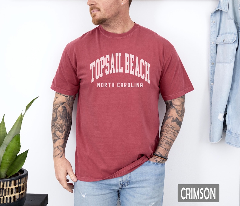 Pu&ograve; includere: T-shirt color cremisi con la scritta bianca "TOPSAIL BEACH NORTH CAROLINA". La maglietta a maniche corte sembra essere realizzata in un materiale morbido e confortevole. La parola "CRIMSON" &egrave; visualizzata in un riquadro rettangolare in basso a destra.