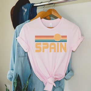 May include: A pink t-shirt with a retro sunset graphic and the word "SPAIN" in a vintage font.
