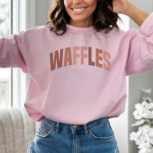 May include: A pink sweatshirt with the word "WAFFLES" printed in a gradient of brown, orange, and pink.