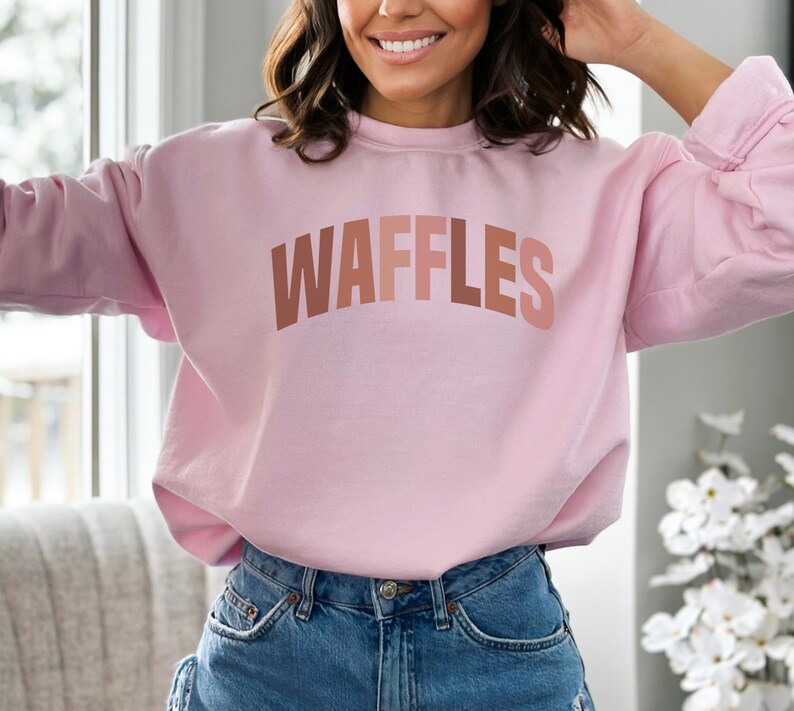 May include: A pink sweatshirt with the word "WAFFLES" printed in a gradient of brown, orange, and pink.