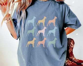 May include: A blue t-shirt featuring a repeating pattern of colorful dog silhouettes. The dogs are in shades of blue, yellow, orange, and green. The shirt is a casual style.