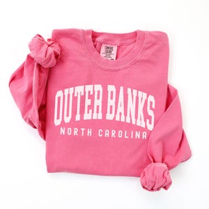 May include: A pink long-sleeved sweatshirt with the words "OUTER BANKS" in large white block letters, and "NORTH CAROLINA" below. The sleeves are cuffed. The sweatshirt is on a white background.