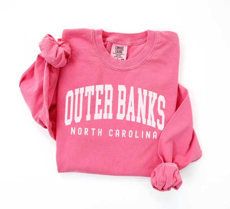 May include: A pink long-sleeved sweatshirt with the words "OUTER BANKS" in large white block letters, and "NORTH CAROLINA" below. The sleeves are cuffed. The sweatshirt is on a white background.