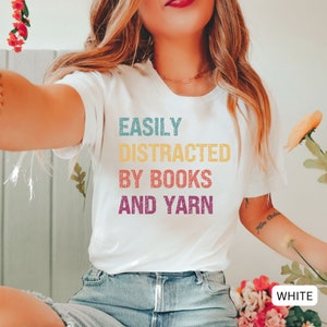 May include: White t-shirt with the text "EASILY DISTRACTED BY BOOKS AND YARN" in colorful, distressed lettering.