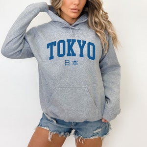 以下が含まれることがあります： グレーのフード付きスウェットシャツに、青い文字で「TOKYO」と、その下に「日本」の日本語が書かれています。