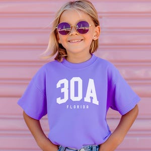 May include: A young person wearing a lavender short-sleeved sweatshirt with the text "30A FLORIDA" in white. The person is also wearing round purple sunglasses with a gold frame. The background is a pink wall.