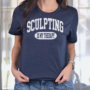 K&ouml;nnte beinhalten: Ein marineblaues T-Shirt mit dem Text "SCULPTING IS MY THERAPY" in wei&szlig;en Blockbuchstaben. Der Text ist in zwei Zeilen angeordnet, wobei "SCULPTING" &uuml;ber "IS MY THERAPY" in einer ovalen Form angeordnet ist. Das Shirt ist aus weichem Material.