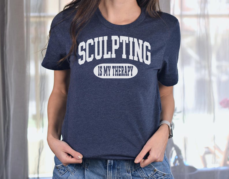 K&ouml;nnte beinhalten: Ein marineblaues T-Shirt mit dem Text "SCULPTING IS MY THERAPY" in wei&szlig;en Blockbuchstaben. Der Text ist in zwei Zeilen angeordnet, wobei "SCULPTING" &uuml;ber "IS MY THERAPY" in einer ovalen Form angeordnet ist. Das Shirt ist aus weichem Material.