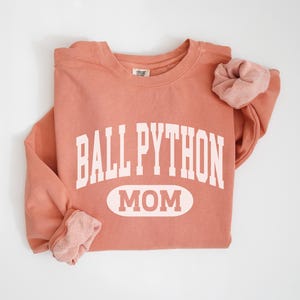 May include: A coral-coloured sweatshirt with the words "BALL PYTHON MOM" printed in white block letters. A matching scrunchie rests on the sleeve. The sweatshirt is folded neatly on a white surface.