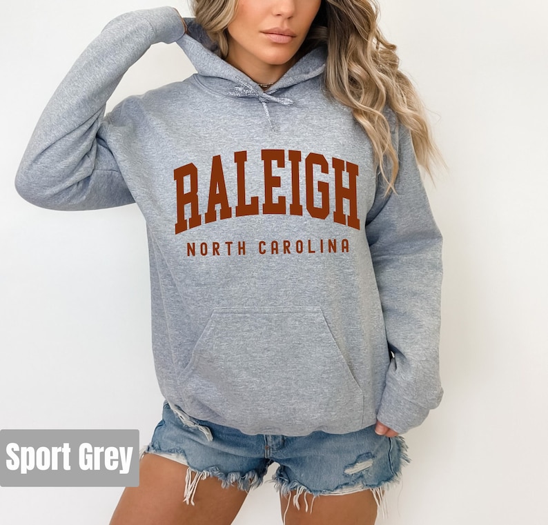 Pode incluir: Uma sudadera com capuz cinza mescla com uma grande estampa marrom que diz "RALEIGH NORTH CAROLINA".