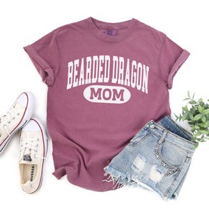 Puede incluir: Una camiseta malva con el texto "BEARDED DRAGON MOM" en blanco, combinada con pantalones cortos de mezclilla y zapatillas blancas. Los pantalones cortos tienen detalles desgastados y adornos. También hay una pequeña planta verde.