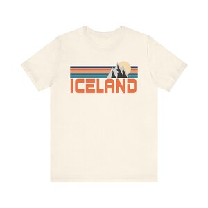 Puede incluir: Una camiseta blanca con un dise&ntilde;o gr&aacute;fico retro que presenta una cordillera, un sol y la palabra "ICELAND" en texto naranja.