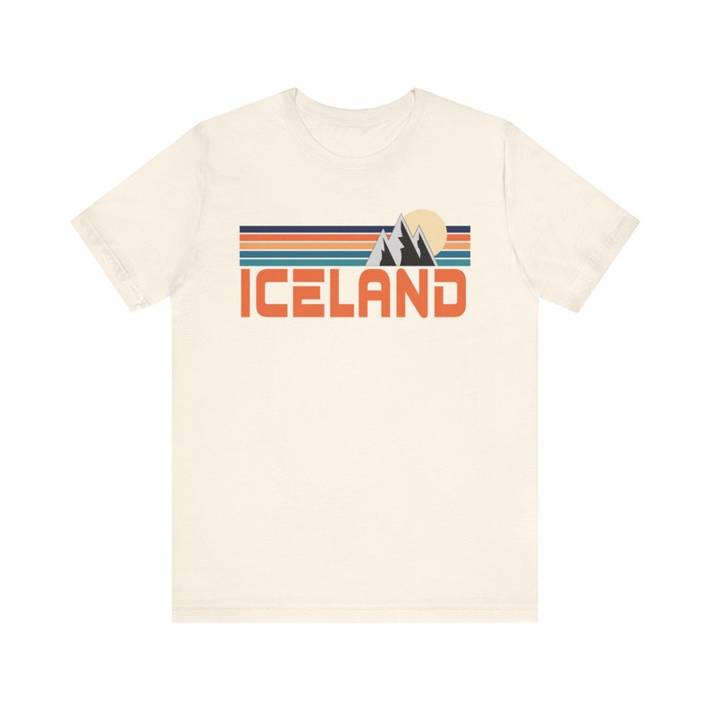 Puede incluir: Una camiseta blanca con un dise&ntilde;o gr&aacute;fico retro que presenta una cordillera, un sol y la palabra "ICELAND" en texto naranja.