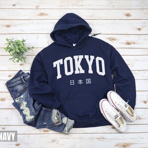 May include: A navy blue hooded sweatshirt with the word "TOKYO" in white lettering and the Japanese characters for "Japan" underneath. The sweatshirt is shown on a white wooden background with a pair of blue jeans and white sneakers.
