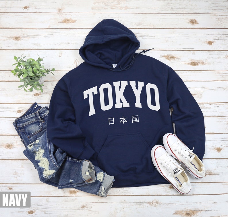 May include: A navy blue hooded sweatshirt with the word "TOKYO" in white lettering and the Japanese characters for "Japan" underneath. The sweatshirt is shown on a white wooden background with a pair of blue jeans and white sneakers.