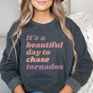 Könnte beinhalten: Ein dunkelgrauer Pullover mit dem Aufdruck "It's a beautiful day to chase tornados" in Rosa und Weiß.