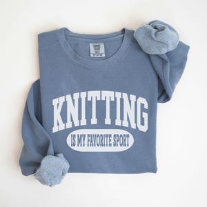 May include: A blue sweatshirt with the words "KNITTING IS MY FAVORITE SPORT" in white. The sleeves are folded, showing the cuffs. The sweatshirt is made of a soft material. The garment is a comfortable fit.