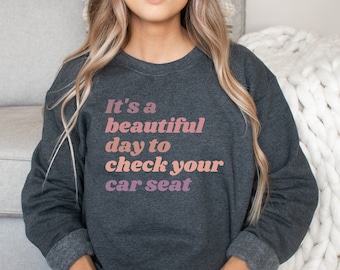 Pode incluir: Uma camisa de moletom cinza escuro com o texto "It's a beautiful day to check your car seat" impresso em rosa e branco.