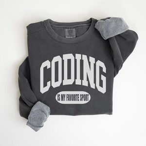 Może przedstawiać: Ciemnoszara bluza z napisem "CODING" dużymi białymi literami na piersi. Poniżej znajduje się napis "IS MY FAVORITE SPORT" w białym owalu. Rękawy zakończone szarym polarem.