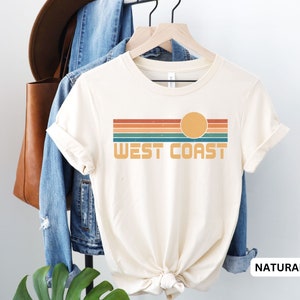 May include: A cream colored t-shirt with a retro-style graphic of a sunset over a striped horizon with the text "West Coast". The shirt is displayed on a hanger with a denim jacket and a brown leather bag in the background. The text "NATURAL" is visible in the lower right corner of the image.