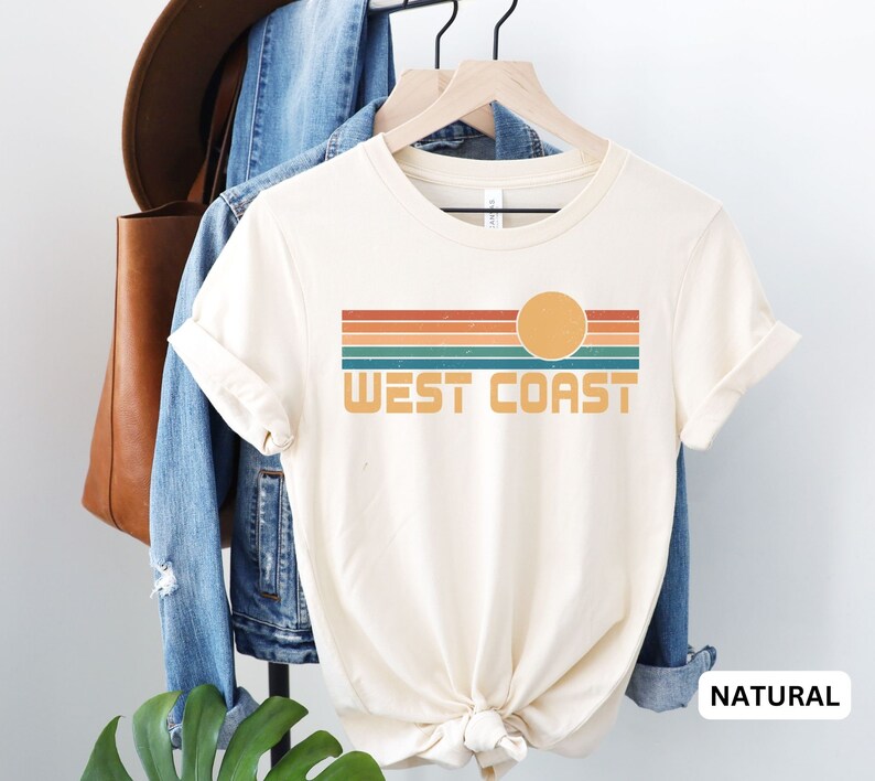 May include: A cream colored t-shirt with a retro-style graphic of a sunset over a striped horizon with the text "West Coast". The shirt is displayed on a hanger with a denim jacket and a brown leather bag in the background. The text "NATURAL" is visible in the lower right corner of the image.