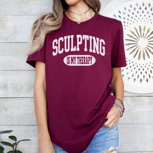 K&ouml;nnte beinhalten: Burgunderfarbenes T-Shirt mit dem wei&szlig;en Text "SCULPTING IS MY THERAPY". Der Text ist mittig auf der Brust platziert. Das Shirt hat kurze &Auml;rmel und scheint aus einem weichen Material zu sein.