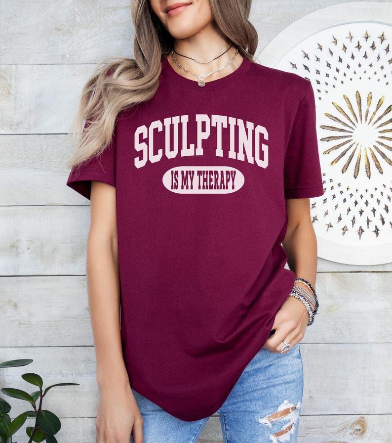 K&ouml;nnte beinhalten: Burgunderfarbenes T-Shirt mit dem wei&szlig;en Text "SCULPTING IS MY THERAPY". Der Text ist mittig auf der Brust platziert. Das Shirt hat kurze &Auml;rmel und scheint aus einem weichen Material zu sein.