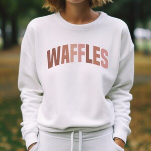 May include: White sweatshirt with a pink and orange gradient text that reads "WAFFLES" on the front.