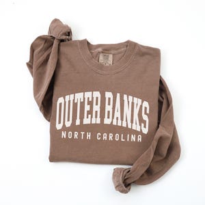May include: A brown long-sleeve shirt with the words "Outer Banks North Carolina" printed in white block letters. The shirt is made of a soft material and has a crew neck. The sleeves are long and cuffed.