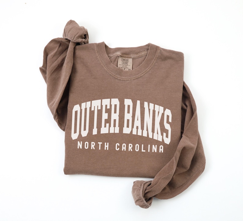 May include: A brown long-sleeve shirt with the words "Outer Banks North Carolina" printed in white block letters. The shirt is made of a soft material and has a crew neck. The sleeves are long and cuffed.