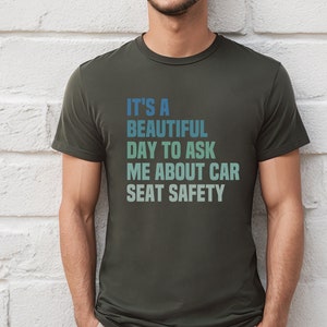 K&ouml;nnte beinhalten: Ein dunkelgr&uuml;nes T-Shirt mit einer wei&szlig;en Grafik, auf der "Es ist ein sch&ouml;ner Tag, um mich nach der Sicherheit von Autositzen zu fragen" steht.