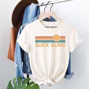 May include: A cream colored t-shirt with a retro-style graphic design featuring a sunset with orange, red, green, and blue stripes and the text "BLOCK ISLAND".