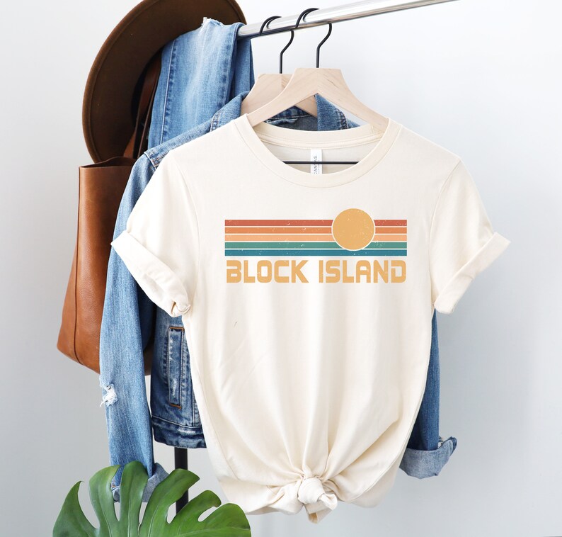 May include: A cream colored t-shirt with a retro-style graphic design featuring a sunset with orange, red, green, and blue stripes and the text "BLOCK ISLAND".