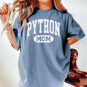 May include: A slate blue t-shirt featuring the white text "PYTHON MOM". The word "MOM" is within a white oval. The t-shirt is a relaxed fit, ideal for casual wear. The image also shows a pair of brown boots.
