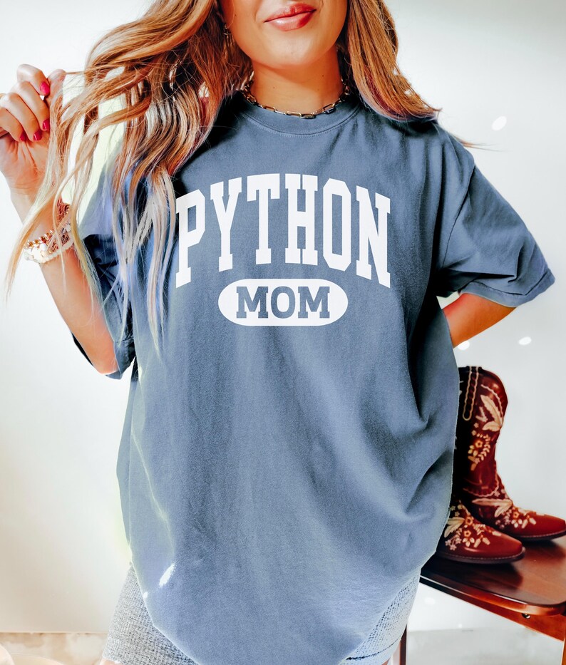 May include: A slate blue t-shirt featuring the white text "PYTHON MOM". The word "MOM" is within a white oval. The t-shirt is a relaxed fit, ideal for casual wear. The image also shows a pair of brown boots.