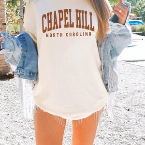 Op de afbeelding: Een wit T-shirt met de tekst "Chapel Hill North Carolina" in bruine letters. Het shirt wordt gedragen door een persoon en is te zien tegen een achtergrond van palmbomen en een blauwe lucht. De tekst "IVORY" staat in de rechterbenedenhoek van de afbeelding.