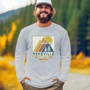 May include: A gray long-sleeve shirt with a vintage-style graphic of mountains, a sun, and trees. The graphic is in a shield shape with the text "Asheville, North Carolina" below it.